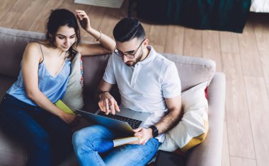 Genç etnik sakallı, gözlüklü, kanepeye oturmuş dizüstü bilgisayarını karıştıran ve yanında oturan genç kadını gösteren serbest bir adamdan.