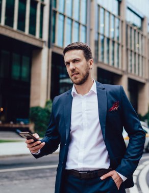 Ciddi bir patron, elinde resmi giysiler, uzaklara bakıyor ve somurtuyor şehir merkezinde yolun kenarında dikiliyor ve akıllı telefonları tarıyor.