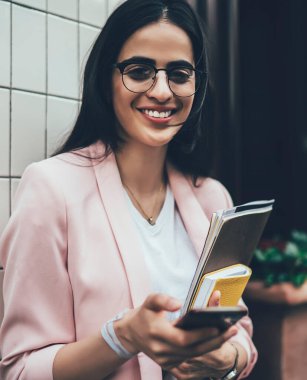 Neşeli, kendine güvenen, gözlüklü ve spor ceketli bir kadın. Elinde kağıt klasör, organizatör ve cep telefonu ile şehrin modern binasının duvarının yanında duruyor.
