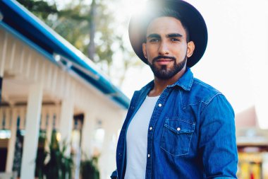 Ciddi sakallı, cepleri dolu, güneşli bir günde şehir sokağının bulanık arka planında dikilirken kameraya bakan bir erkek.