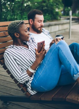 Birbirine aşık çok ırklı sakin çiftin yan görüntüsü ahşap park şezlongunda zevkle dinleniyor ve cep telefonu kullanıyor.
