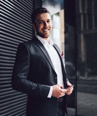 Koyu takım elbiseli, cep telefonlu neşeli bir işadamı metal duvarın yanında duruyor ve şehir merkezinde kameraya bakıyor.