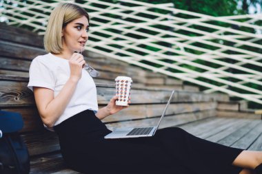 Resmi kıyafetler giymiş, gözlüklü, ev yapımı içkili, dizüstü bilgisayarlı, tahta merdivenlerde oturan ve gözlerini kaçıran düşünceli yetişkin bir kadının yan görüntüsü.