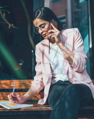 Aşağıdaki mutlu yetişkin serbest çalışan, gözlüklü, sıcak içecekle resmi giyinmiş, telefon görüşmesi yapan, not defterine yazan ve caddede bulanık bir şekilde oturmakta olan.