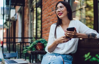 Yüzünde sevimli bir gülümsemeyle mutlu bir Latin kadın elinde dijital cep telefonu cihazıyla ve boş zamanlarında internetteki telefon konuşmaları için seviniyor. Akıllı telefonlu neşeli bir bayan milenyumu kafenin terasında oturuyor.