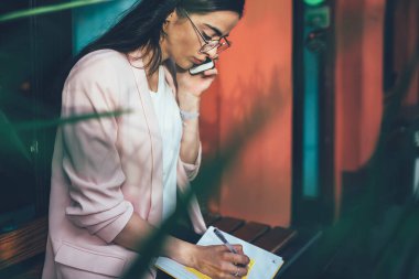 Gözlüklü ve zarif elbiseli ciddi yönetici bir kadının ahşap bankta otururken not defterine yazması ve telefon konuşması yapması.