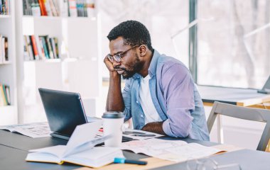 Gözlüklü, tişörtlü, dizüstü bilgisayarlı bir Afro-Amerikan adam gözlüklü, not defterleriyle dolu bir masada oturuyor.
