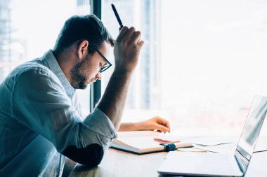 Mavi gömlekli, gözlüklü, aklı başında bir entelektüel. Pencere karşısında dizüstü bilgisayarla masada oturan not defterini kontrol ediyor.