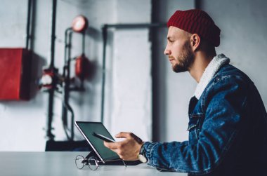 Genç dalgın sakallı, stil sahibi serbest stil sahibi, sıcak mavi kot ceket giyen, kırmızı şapkalı iş yerindeki aletleri kullanan