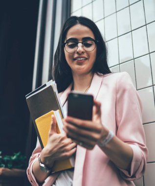 Elinde kağıtlar ve günlükle bina duvarının yanında duran ve internette cep telefonundan haber okuyan başarılı, modern meşgul bir kadından.