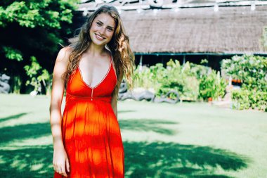 Koyu saçlı, kıvırcık, neşeli, dişlek, kırmızı elbiseli, neşeli bir kadın bulutsuz bir günde yeşil parkta dinleniyor.