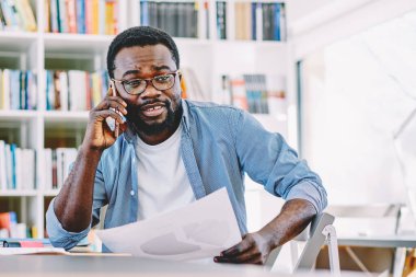 Gözlüklü, mavi tişörtlü, cep telefonlu, elinde belgeyle masada oturan zeki Afro-Amerikan bir adam.