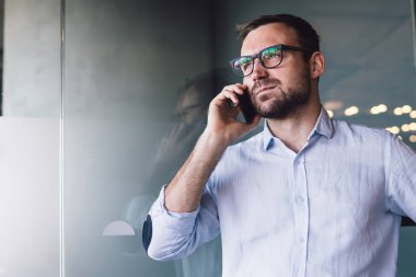 Mavi gömlekli ve mavi pantolonlu ciddi sorunlu bir erkek camdan kapının karşısındaki ofiste cep telefonundan arama yapıyor.