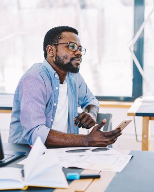 Orta yaşlı Afrikalı Amerikalı adam çalışırken bilgisayar ve kağıtlarla masada otururken akıllı telefonu elinde tutuyor ve düşünüyor.