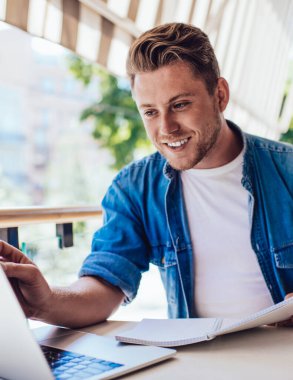 Açık kahvede dizüstü bilgisayar ve ders kitabı kullanırken gülen gündelik kıyafet uzmanı sarışın bir erkek.