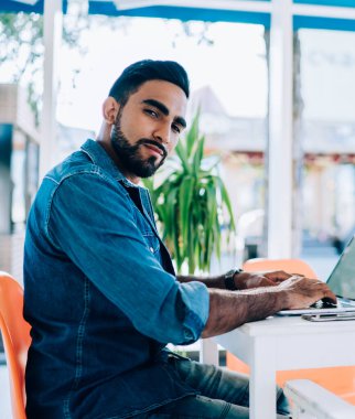 Dizüstü bilgisayar kullanarak yaz kafeteryasında oturup kameralara bakarak proje üzerinde çalışan serbest giyinen sakin etnik bir vatandaş.