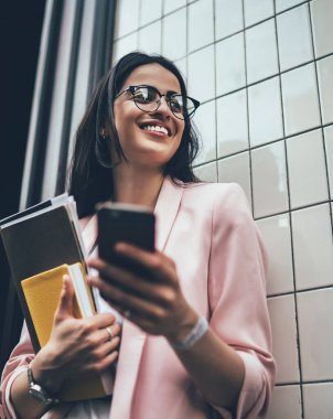 Gözlüklü ve spor ceketli neşeli bir kadının elinde kağıt klasör ve günlükle bina duvarının yanında dururken çok heyecanlı bir şekilde cep telefonu kullanması ve gülümsemesi.