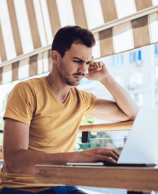 Güneşli bir günde, kafenin açık terasında dizüstü bilgisayarında problemle uğraşan şaşkın, serbest çalışan erkek oyunculardan.