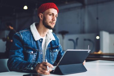 Yoğunlaşmış, şık, sakallı, erkek serbest çalışan, ofiste çalışan, kalın paltolu ve şapkalı, bulanık arkaplanlı tabletli bir adam.