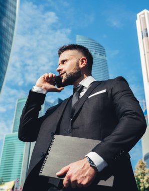 Başarılı iş adamlarından takım elbiseli, telefonla konuşan, dosyalar taşıyan ve modern ofis binalarını arka planda tutan biri.