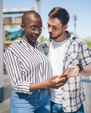 Elinde cep telefonu ve sakallı adam olan konsantre olmuş genç siyahi kadın, sokak sanat galerisinde yan yana dururken aleti işaret ediyor.