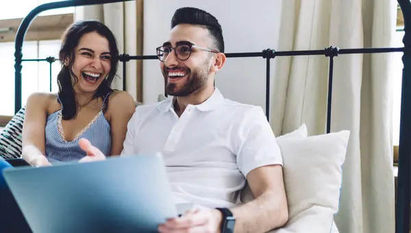 Bearded adult man in glasses and casual clothes holding laptop on knees and pointing with hand on screen while sitting on bed and laughing with pretty woman in light bedroom