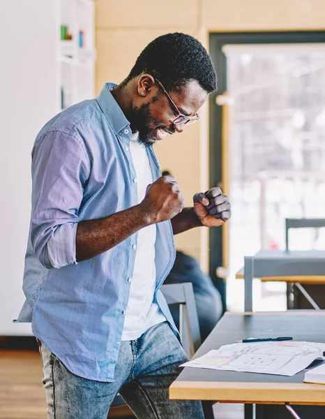 Side view of delighted African American man in casual outfit clenching fists and celebrating success after finishing blueprint in modern workplace