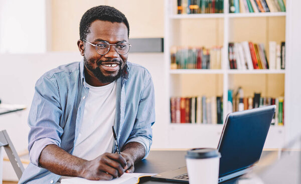 Happy successful African American man in glasses and casual clothes taking notes while sitting with laptop in light design office