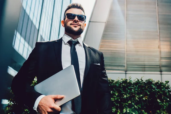 From below charismatic confident executive man in sunglasses and elegant black suit with laptop looking away and smiling while standing on street and enjoying own success against contemporary high