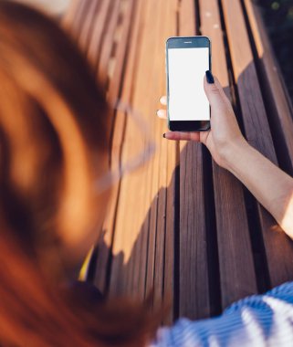 Kırpılmış, kızıl saçlı, sıradan giyinmiş, telefon kullanarak sosyal medyada arkadaşlarıyla sohbet eden, ahşap bankta oturan kadınların arka plan görüntüsü.