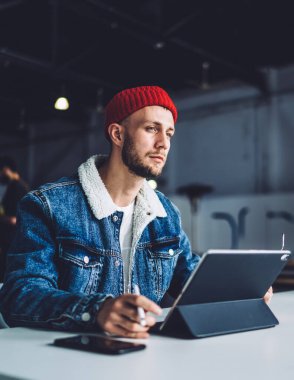Yoğunlaşmış, şık, sakallı, erkek serbest çalışan, ofiste çalışan, kalın paltolu ve şapkalı, bulanık arkaplanlı tabletli bir adam.