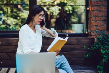 Klasik gözlüklü İspanyol serbest yazar, uluslararası cep telefonu konuşmaları sırasında proje detaylarını tartışmak için not defterinden bilgi kontrol ediyor, milenyum kadını hareketlilik bağlantısı kullanıyor