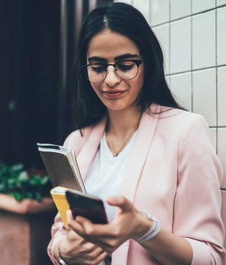 Gözlüklü ve spor ceketli modern meşgul kadın elinde kağıtlar ve günlükle belediye binasının duvarının yanında dururken müşteriden acil e-posta okuyor.