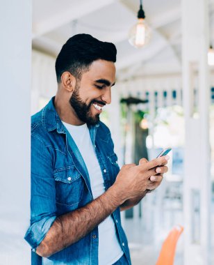 Gündüz kıyafetleri içinde gülümseyen, iyimser etnik erkek cep telefonu kullanıyor sosyal medyada arkadaşlarıyla sohbet ediyor güneşli bir günde.