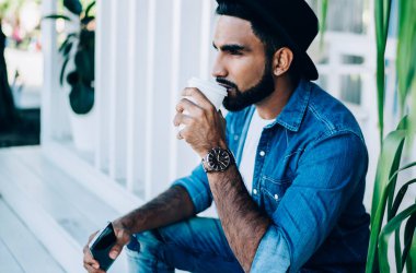 Kot pantolon giymiş, şapkalı, sıcak kahve içen ve şehir binasının yakınındaki akıllı telefonuyla başka yöne bakan düşünceli, sakallı İspanyol adamın yan görüntüsü.