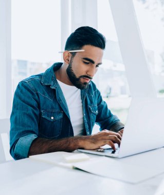 Çalışkan, düşünceli, günlük giysiler içinde, kulağının arkasında kalemle ekranda gezinen ve bilgisayarla etkileşime giren İspanyol asıllı serbest yazar açık havada beyaz terasta elinde not defteriyle otururken.