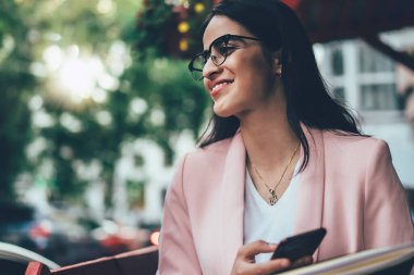 Havalı giyinmiş, gözlüklü, sosyal medyada sörf yapan, kafede vakit geçiren heyecanlı etnik gülümseyen kadın.