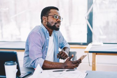 Orta yaşlı Afrikalı Amerikalı adam çalışırken bilgisayar ve kağıtlarla masada otururken akıllı telefonu elinde tutuyor ve düşünüyor.