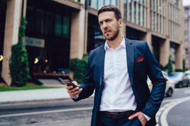 Ciddi bir patron, elinde resmi giysiler, uzaklara bakıyor ve somurtuyor şehir merkezinde yolun kenarında dikiliyor ve akıllı telefonları tarıyor.