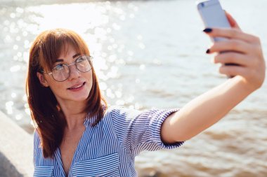 Şehvetli gülümseyen kızıl saçlı, günlük kıyafetler ve gözlükler takan akıllı telefon kullanan, güneşli güneşin altında selfie çeken genç bir kadın.
