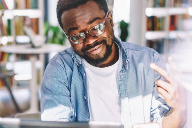 Gözlüklü ve günlük kıyafetleriyle mutlu siyah erkek tasarımcı cep telefonuyla selfie çekiyor ve hafif sanat ofisinde çalışırken zafer jesti yapıyor.