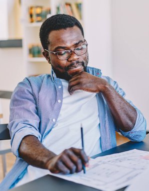 Sıradan giyinmiş, gözlüklü zeki Afro-Amerikan erkek. Masada oturup ofiste proje üzerinde çalışırken planlar üzerinde kafa yoruyor.