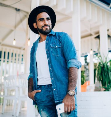 Akıllı telefonlu pozitif sakallı İspanyol adam. Elini cebine atıyor ve sokakta rahat bir restoranın önünde dikiliyor.