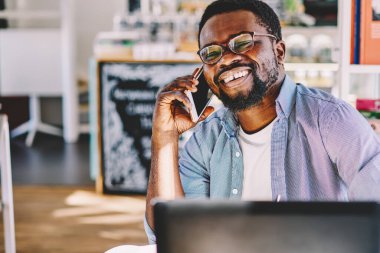 Gözlüklü ve günlük kıyafetleriyle neşeli Afrikalı erkek. Bilgisayarlı masada oturmuş güncel ofis kağıtlarıyla sohbet ediyor.