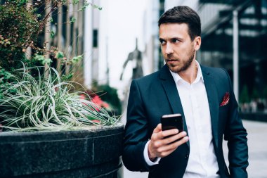 Takım elbiseli başarılı bir genç, partneriyle telefonda iletişim kuruyor. İş yerinin caddesinde oturup gözlerini kaçırıyor.