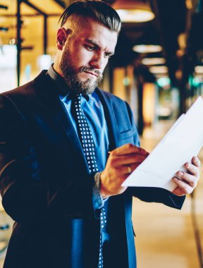 Modern cam ofis koridorunda dikilirken takım elbiseli başarılı sakallı erkek menajer iş evraklarını imzalıyor ve inceliyor.