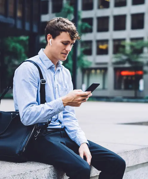 Young male worker in formal shirt sitting with leather bag on concrete fence and messaging on phone on background of New York