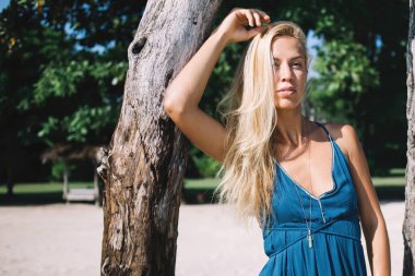 Sarışın, genç, düşünceli, mavi elbiseli bir kadın güneşli bir günde ağaçların arasında dikiliyor ve başka tarafa bakıyor.