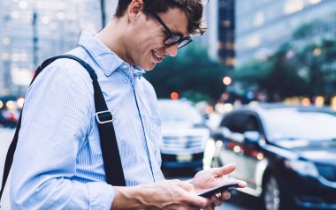 Tişörtlü ve gözlüklü genç erkek çalışanın New York caddesinde durup telefonda mesajlaşırken gülümsemesi.