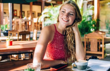 Oldukça neşeli, kırmızı elbiseli yetişkin bir turist. Ahşap masada tek başına oturmuş kitap, akıllı telefon ve içecek içerek kafasını çeviriyor.